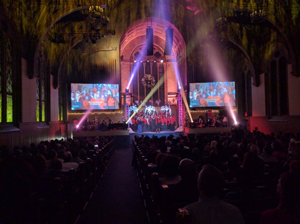 Audience at a stage show in a large hall with screens, stage lighting, and performers.