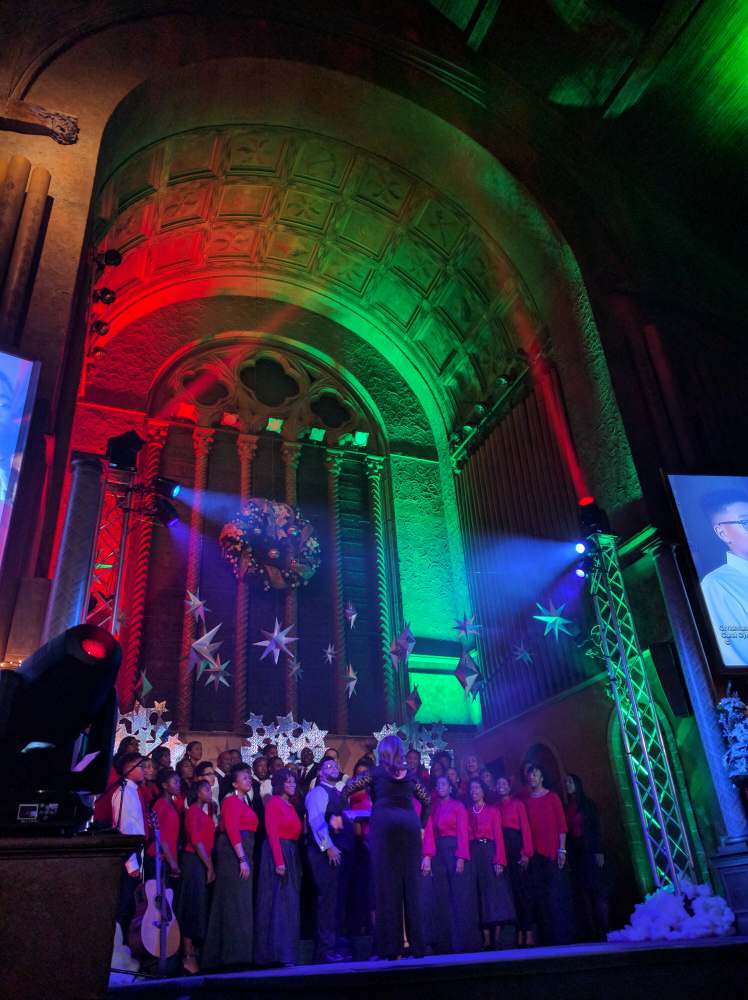 Choir singing on stage in a church decorated with Christmas lights; red and green lighting on arch.