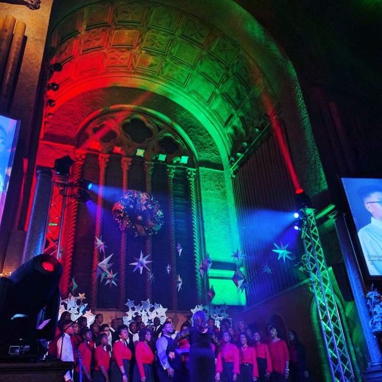 Choir performing on stage in a decorated church with red, green, and blue lighting.