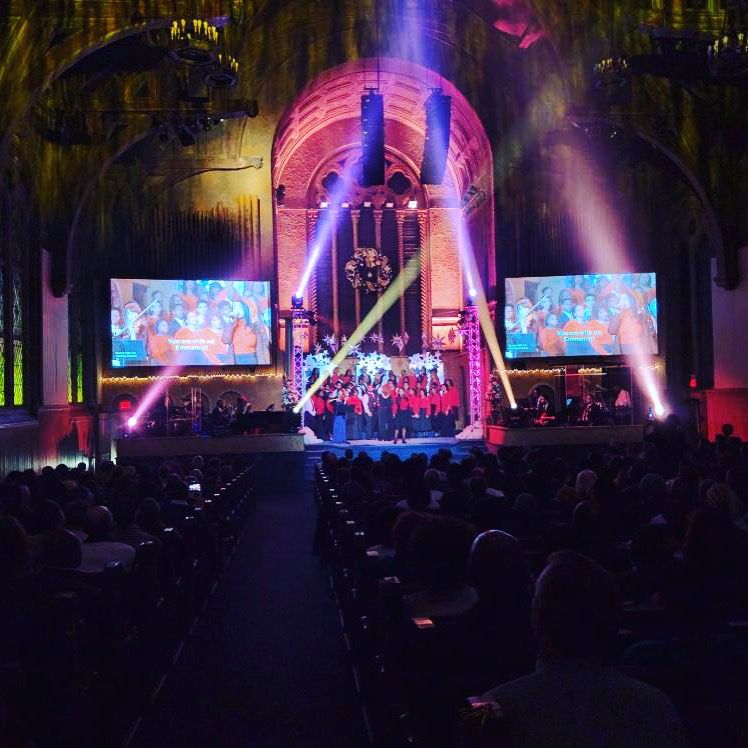 Choir performing on stage in a large auditorium with colorful stage lighting, and two screens with video.