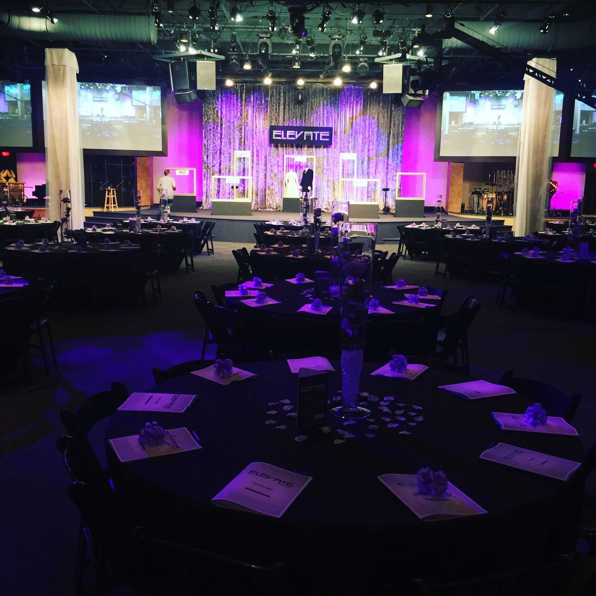 Event hall with round tables set for a gathering, stage in the background with purple lighting.