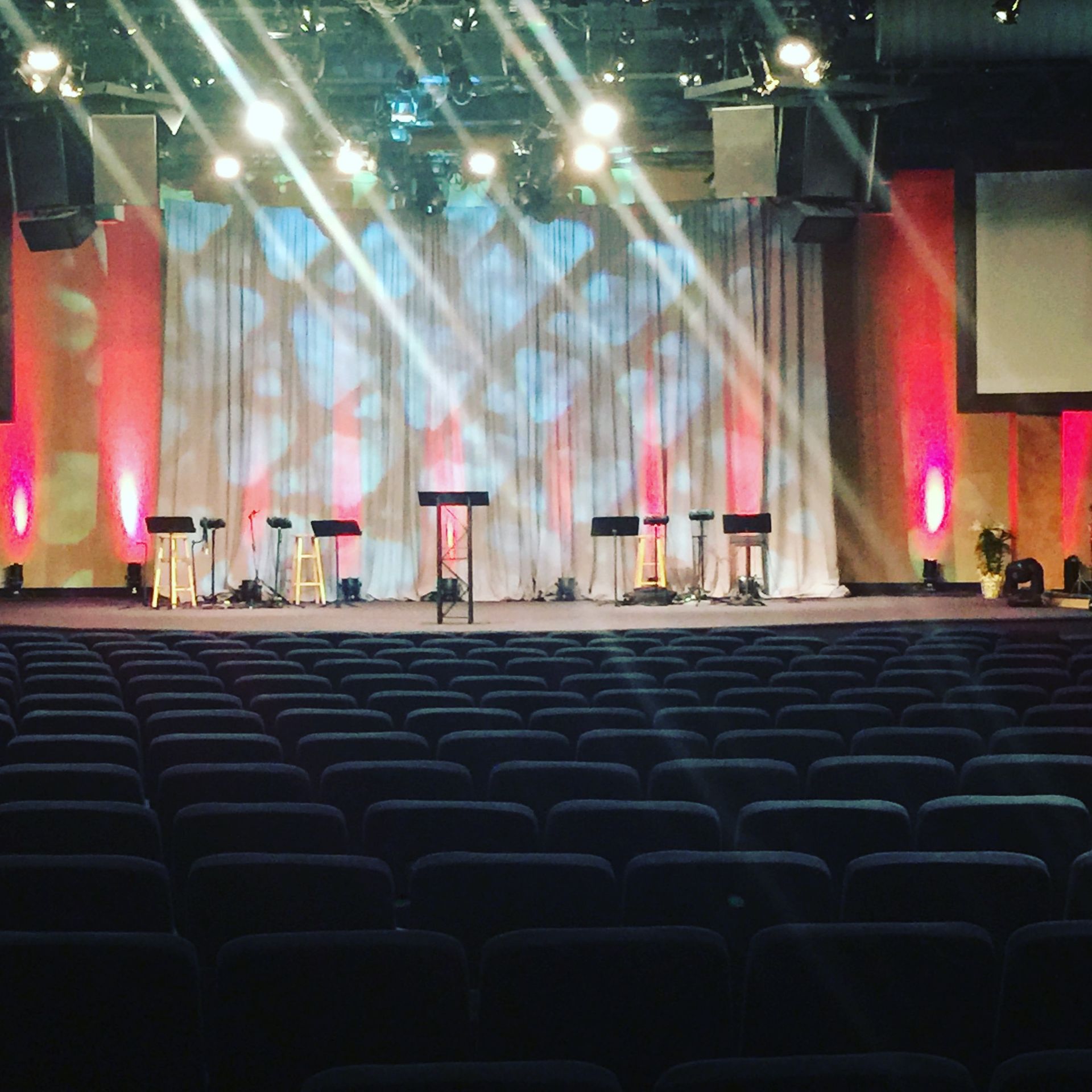 Auditorium with stage setup: rows of seats facing a stage with lights, microphones, and a backdrop.