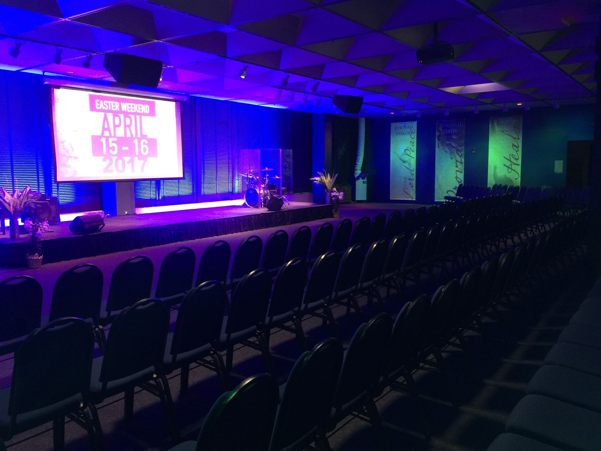 Empty auditorium stage with rows of chairs, lit in blue and green for an event.