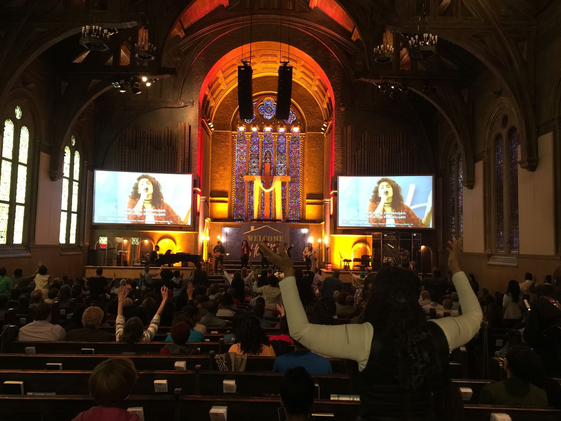 People in a church with raised hands, watching two screens featuring a woman singing. Stage lit with orange and gold.