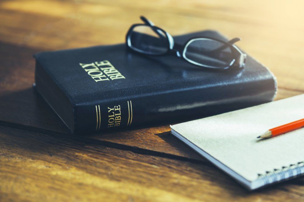 Bible with eyeglasses, notepad, and pencil on a wooden surface.