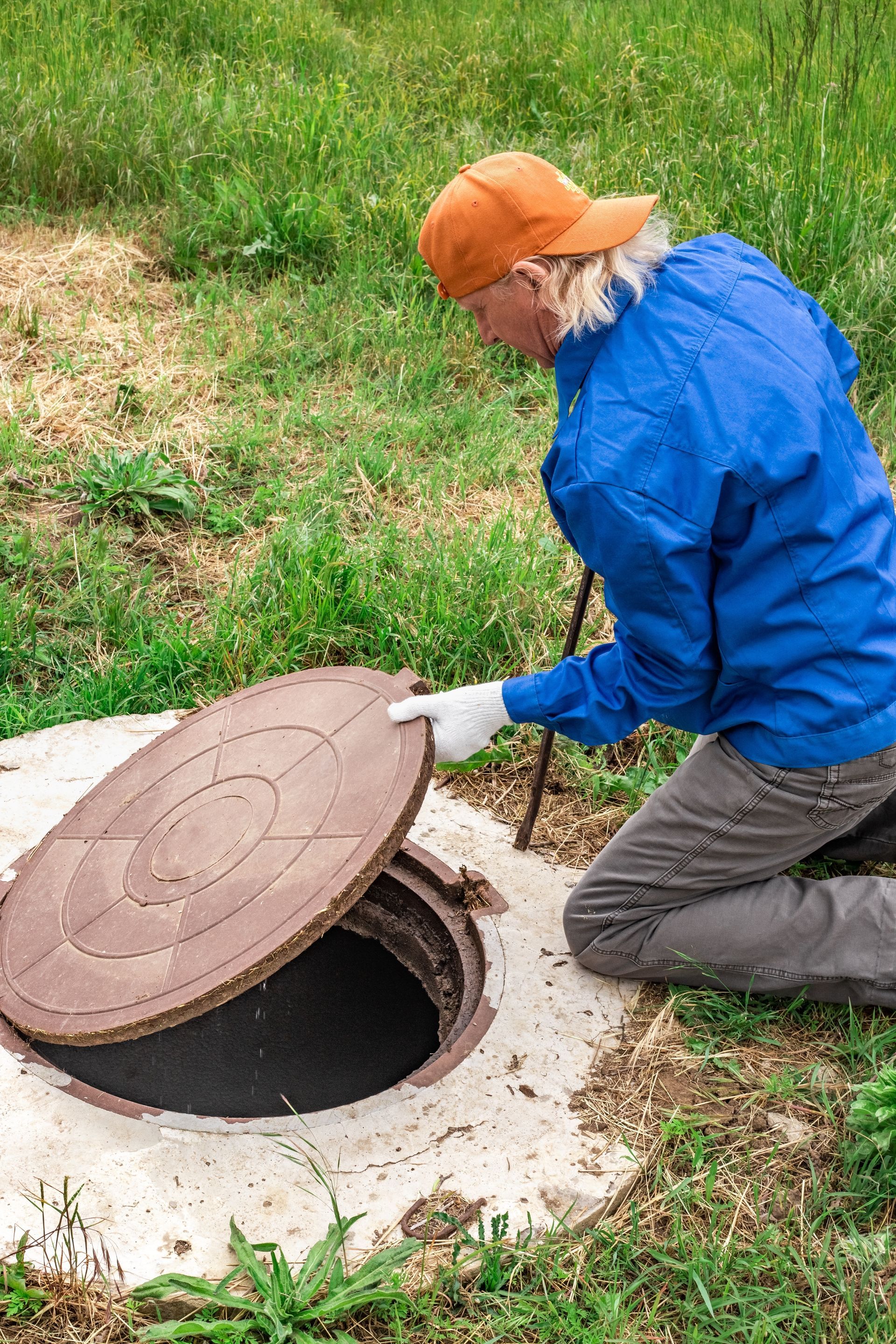 Worker in blue jacket and orange cap lifting a manhole cover on grass