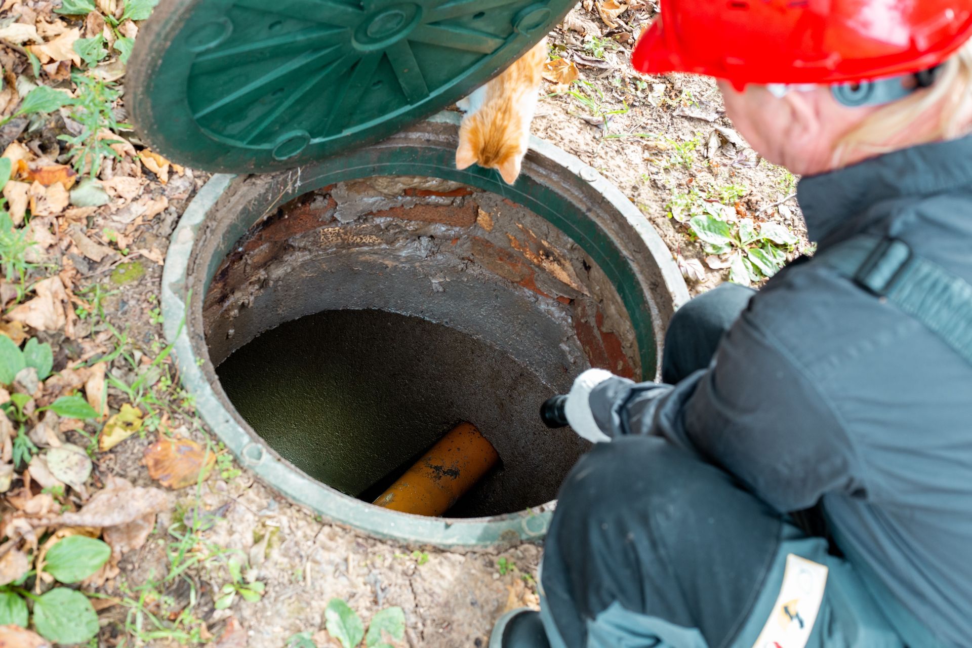 Worker in red hard hat inspecting an open manhole with a lifted green cover.