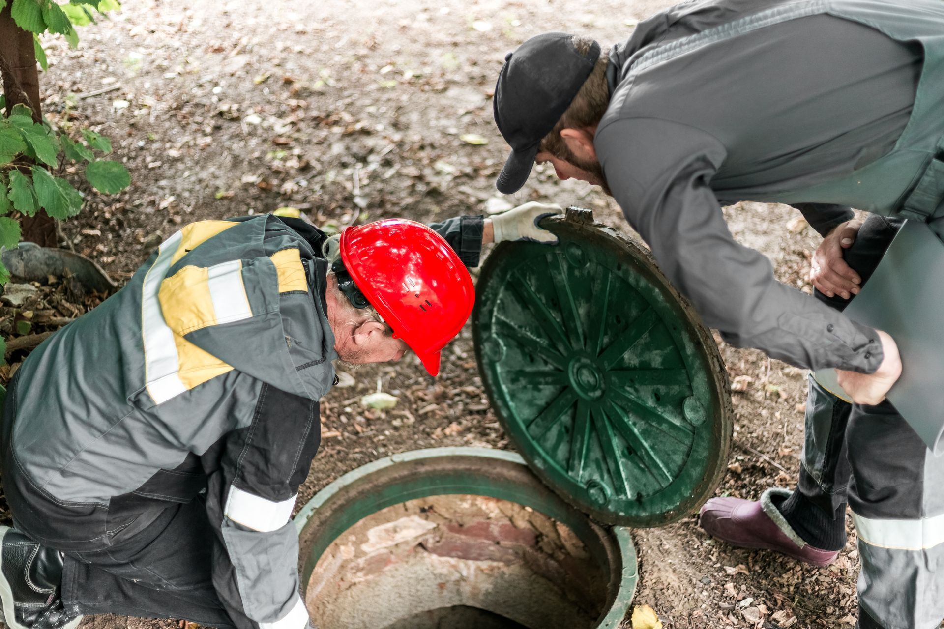 Two workers in hard hats inspect an open green manhole cover outdoors.