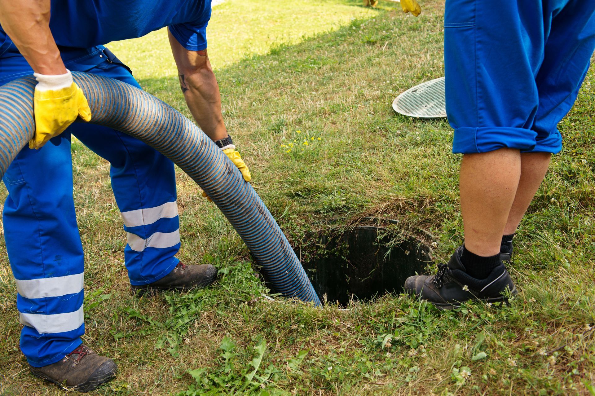 Two workers in blue uniforms use a hose to clear a clogged manhole in a grassy area.