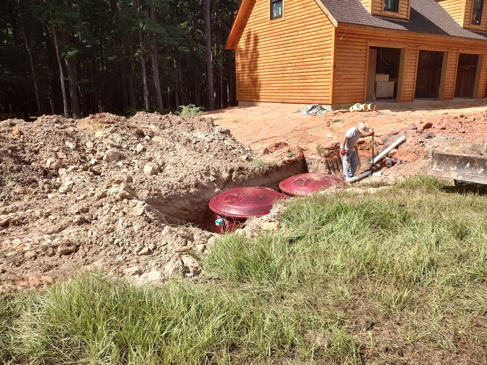 Worker beside muddy trench and red utility covers near a log cabin in a wooded area