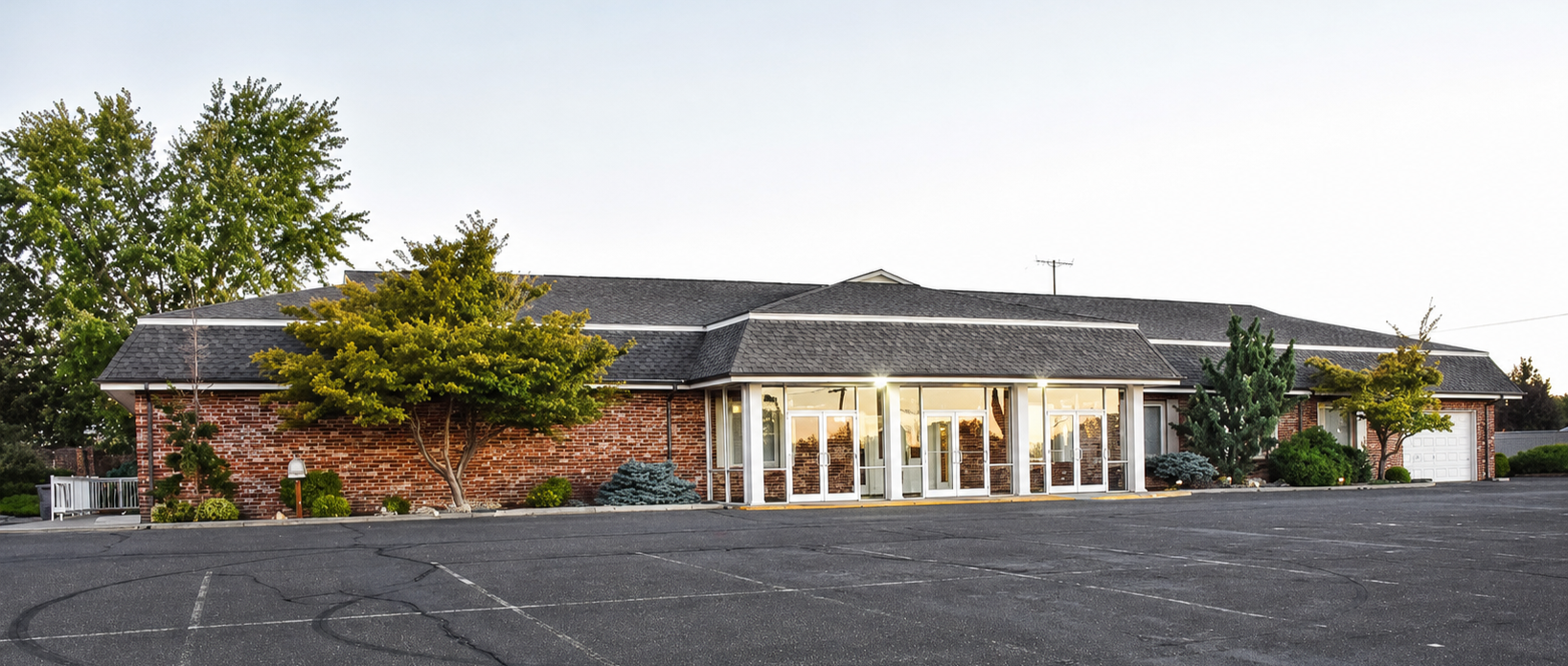 Low brick building with a glass entrance and dark roof, viewed across an empty parking lot
