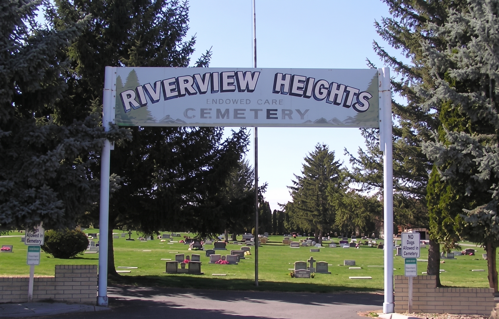Entrance arch reading “Riverview Heights Cemetery” above a grassy cemetery with trees and headstones.