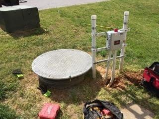 Circular septic tank with a metal lid, PVC pipe structure, electrical box, and tools on a grassy lawn.