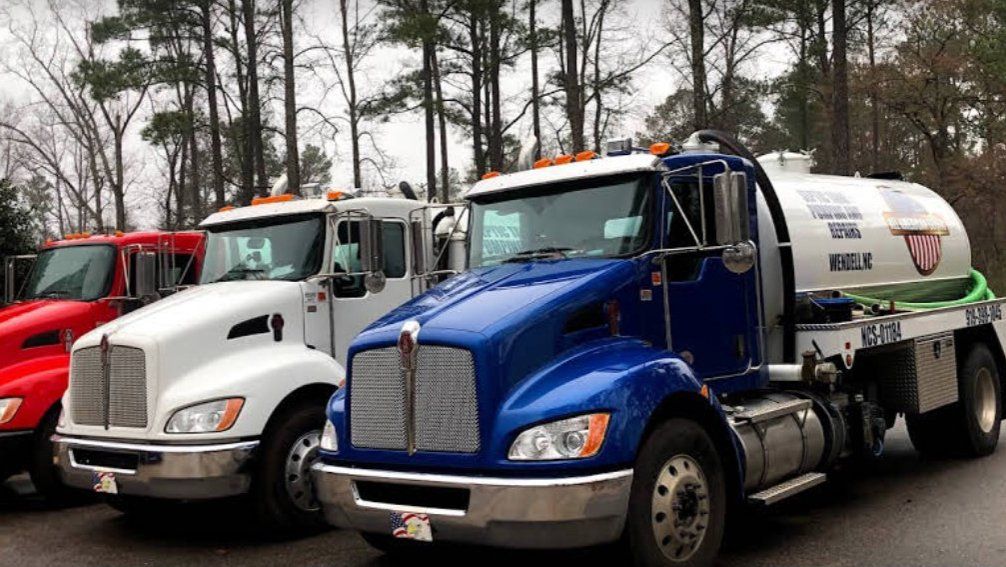 Three septic service trucks parked outside, red, white, and blue, under trees.