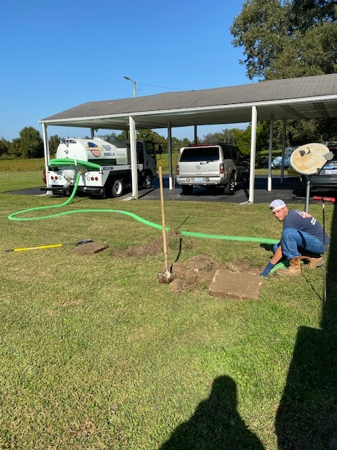 A septic tank truck pumping a septic tank on a lawn; a worker kneels near the open tank.