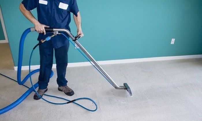 Person in blue uniform cleaning carpet with a steam cleaner in a room with teal walls.