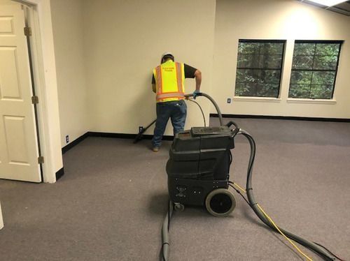 A worker in a safety vest cleans carpet in a room with a large carpet cleaning machine.