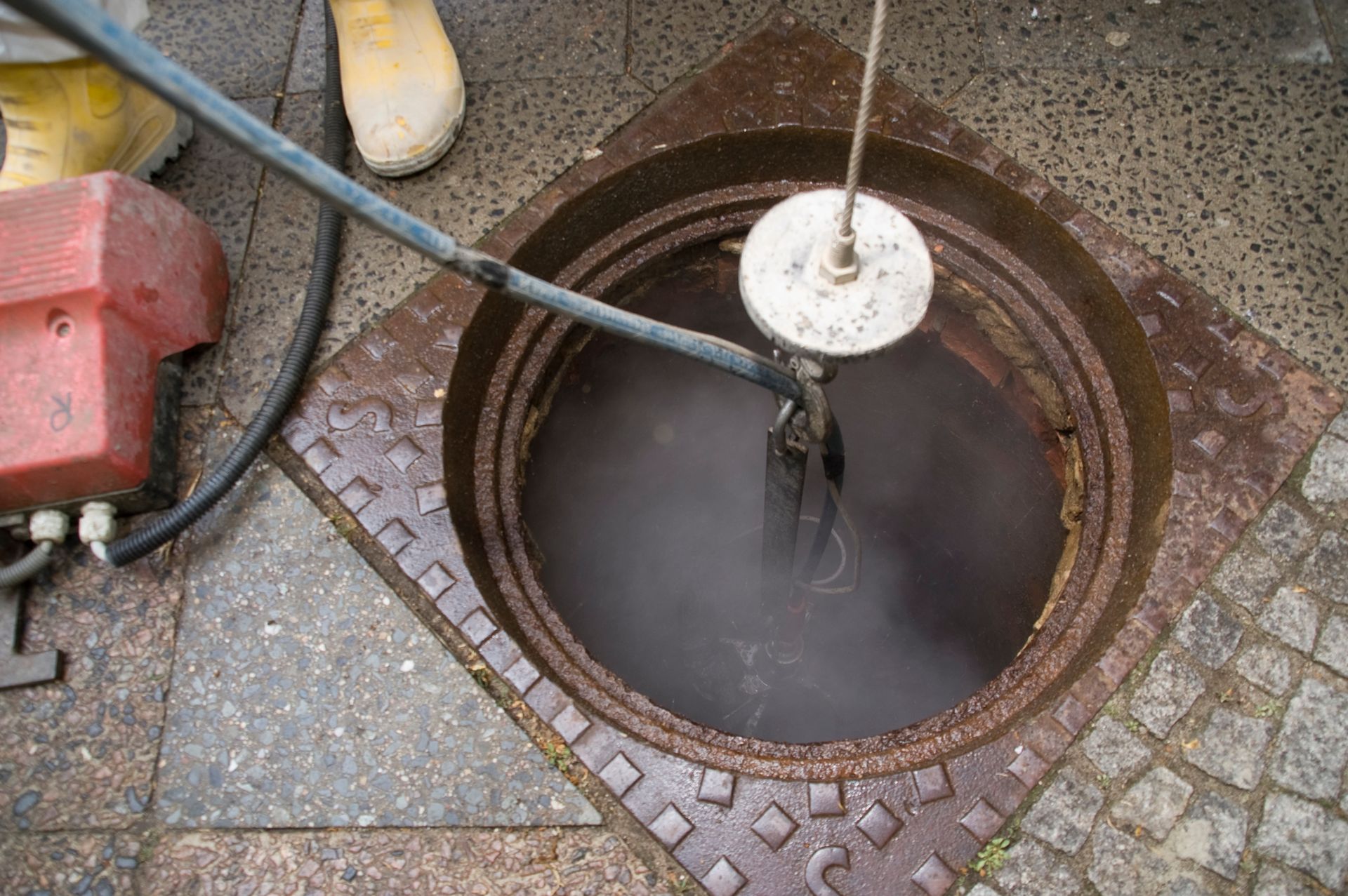 Manhole open on a sidewalk, with a tool inside the dark water. Someone wearing yellow boots.
