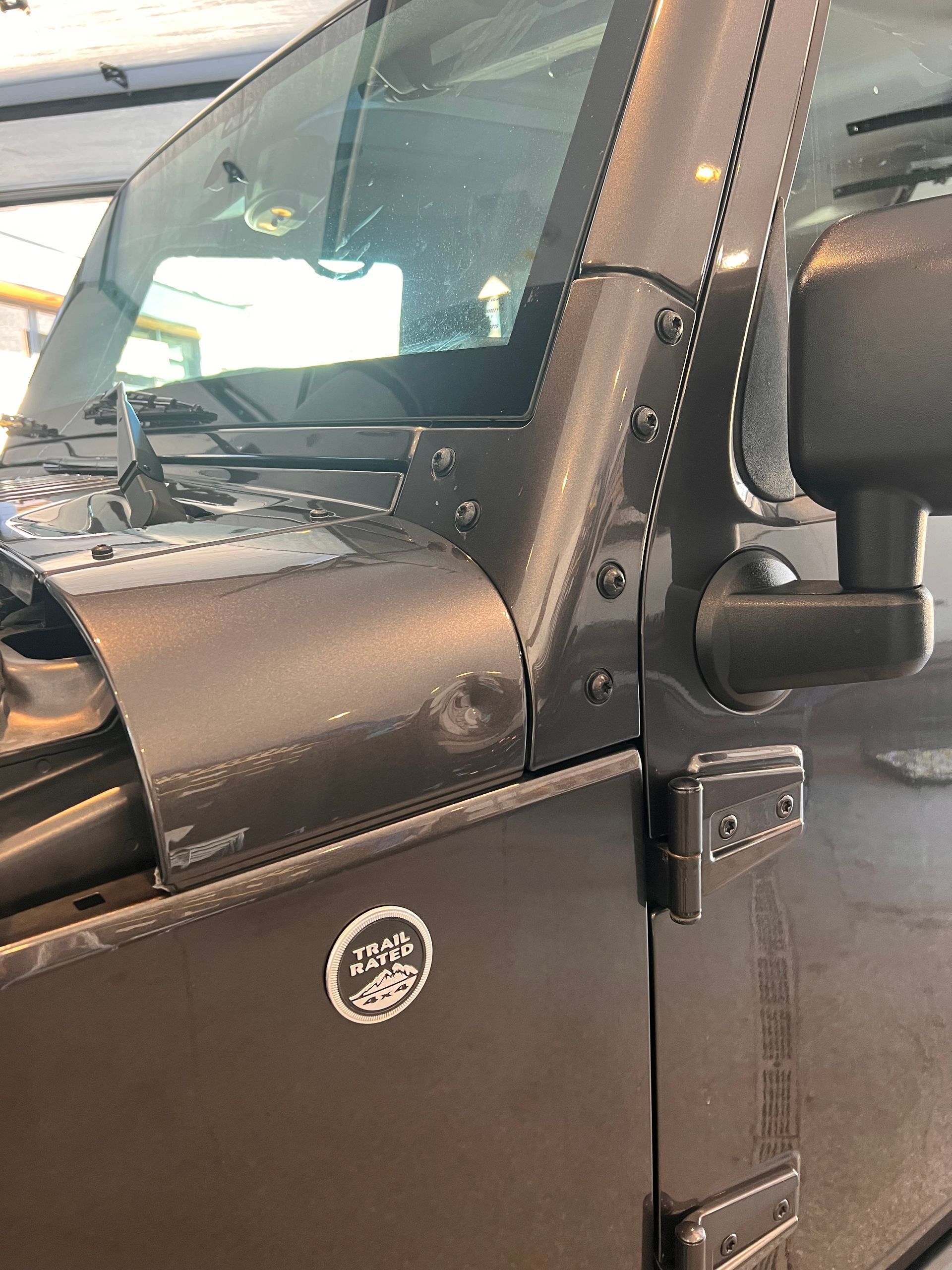 A close-up of a charcoal-colored Jeep exterior, focusing on the windshield frame, driver-side mirror, and door hinge.