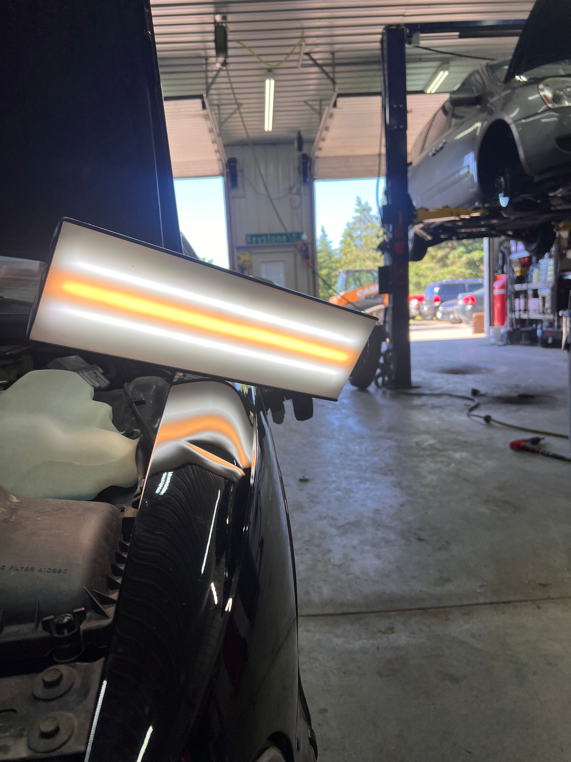 A PDR light board reflects on a dented black car fender in a repair shop with a car on a lift in the background.