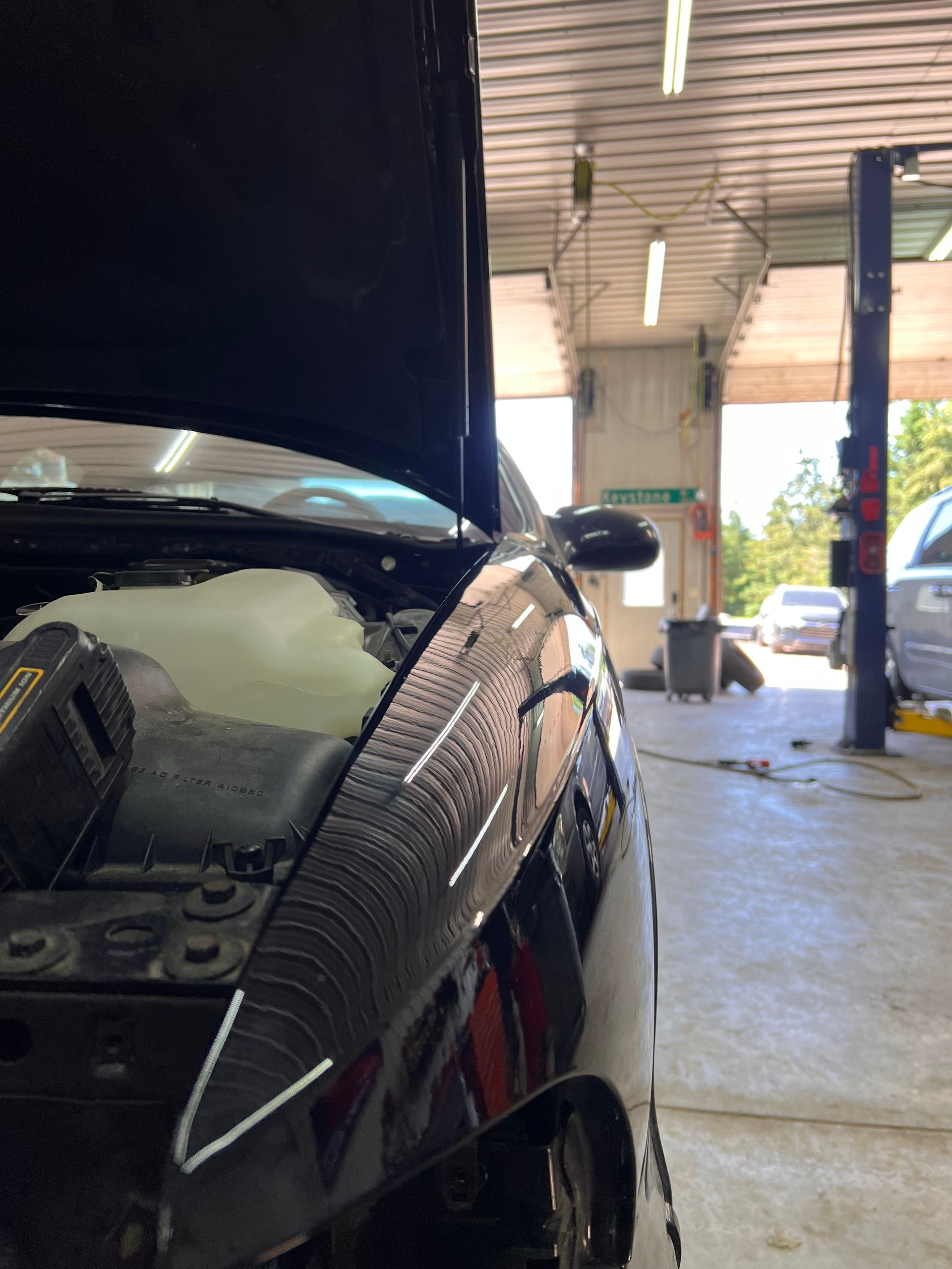 A black car with an open hood is parked in a repair garage, with a view through the shop bay to the outside.