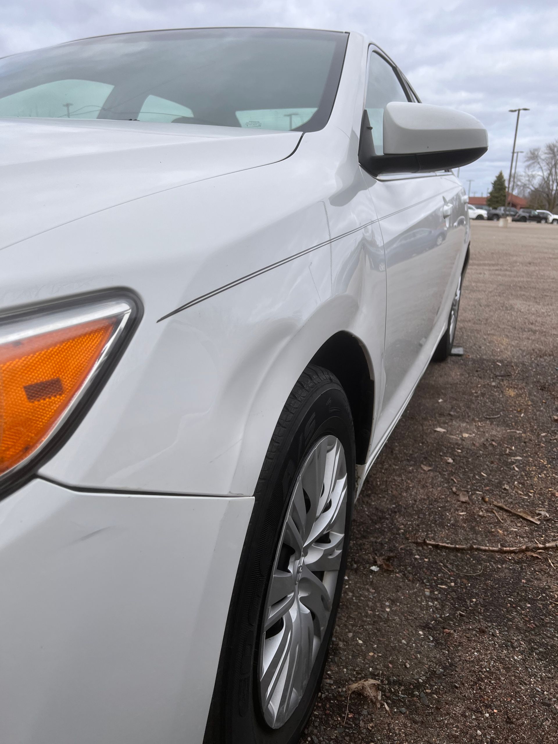A side view of a white car parked outdoors, showing a dent on the front passenger-side fender.
