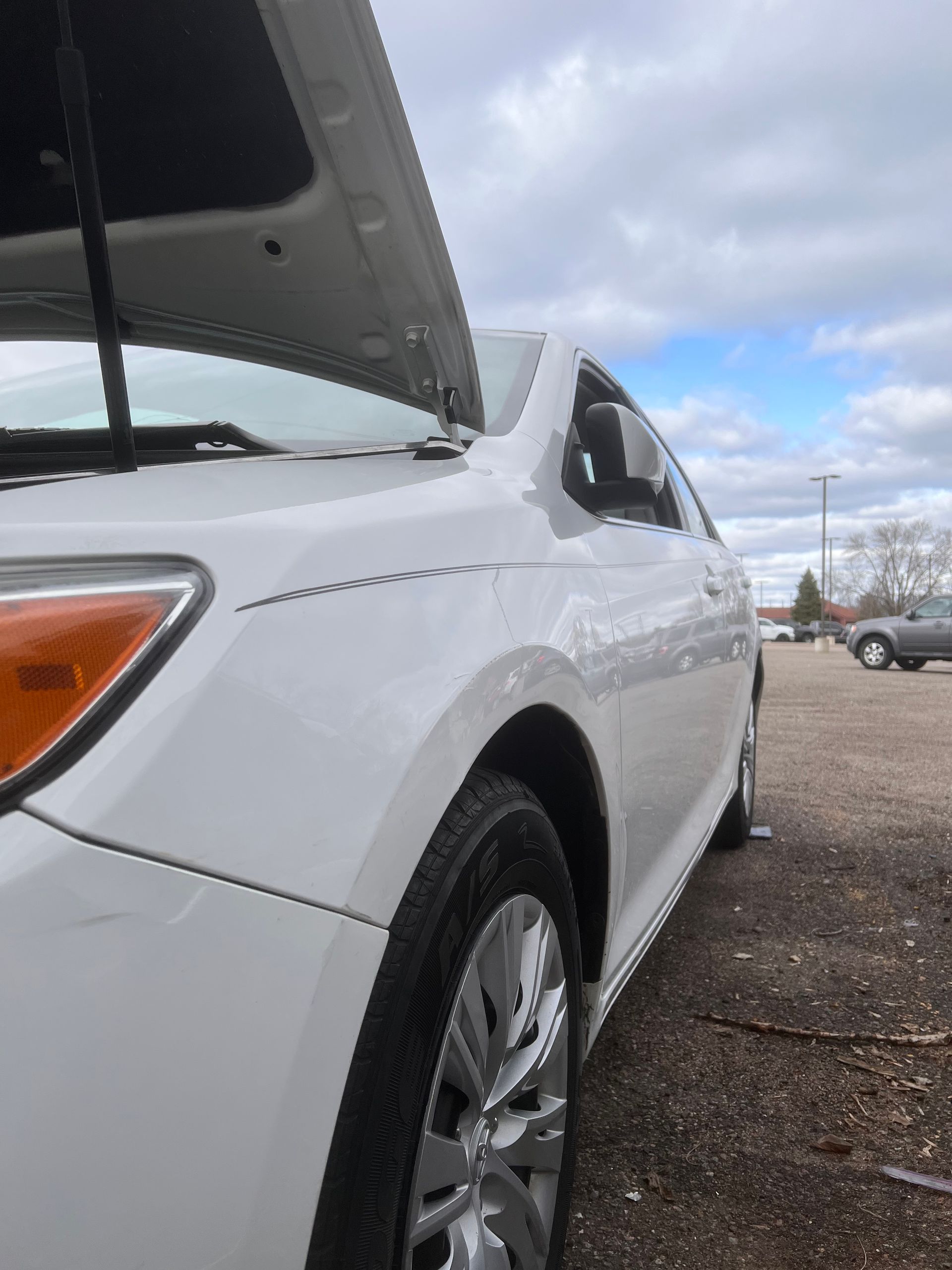 A side view of a white car parked outdoors with its hood open, showing the front passenger side panel and tire.