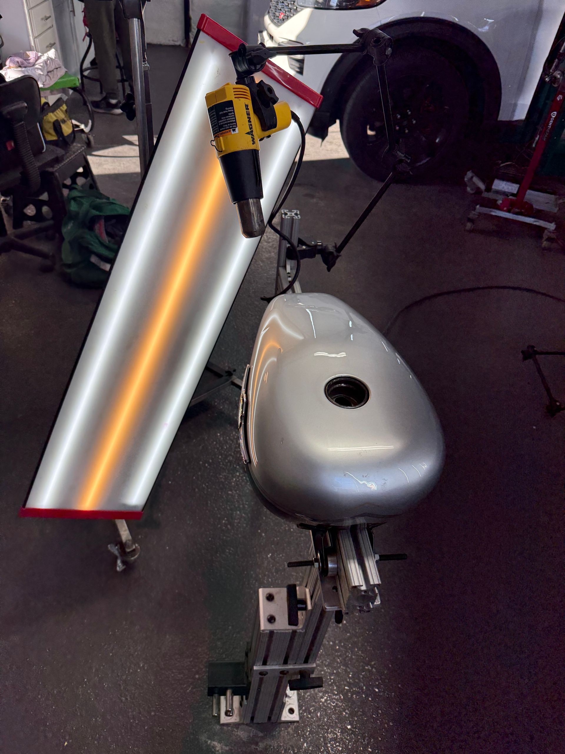 A silver motorcycle gas tank mounted on a stand in a workshop, next to a reflective light board and a yellow heat gun.