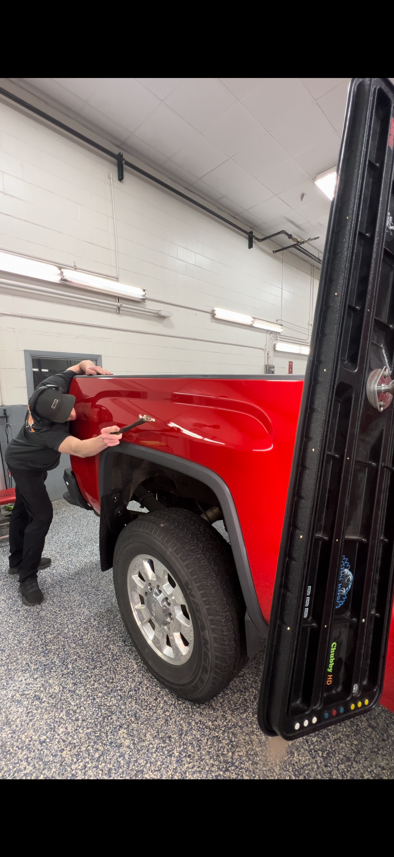 A person in a garage buffs the red rear fender of a pickup truck with a polishing tool.