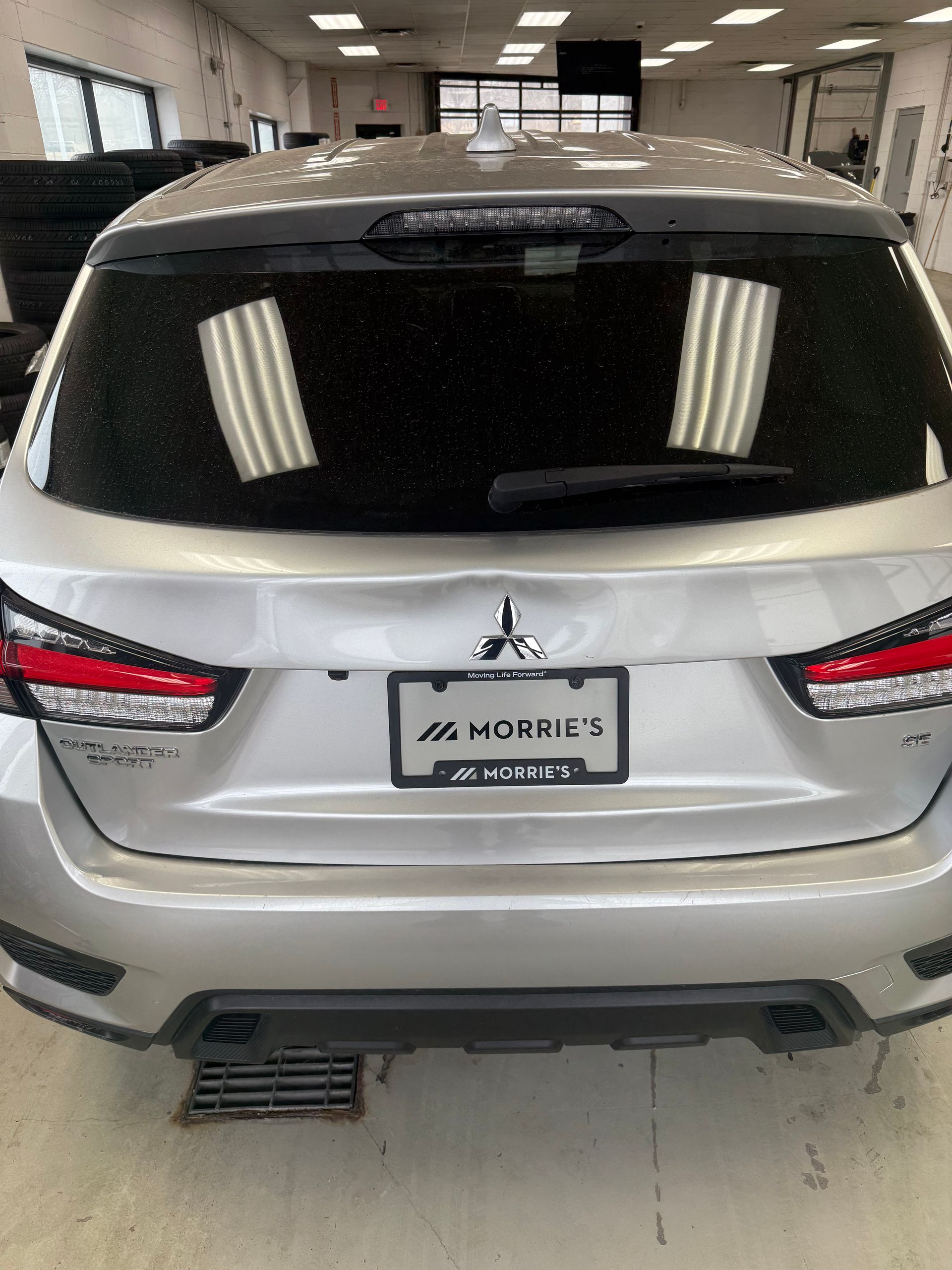 Silver Mitsubishi SUV in an auto shop with significant denting and damage to the center of the rear liftgate.