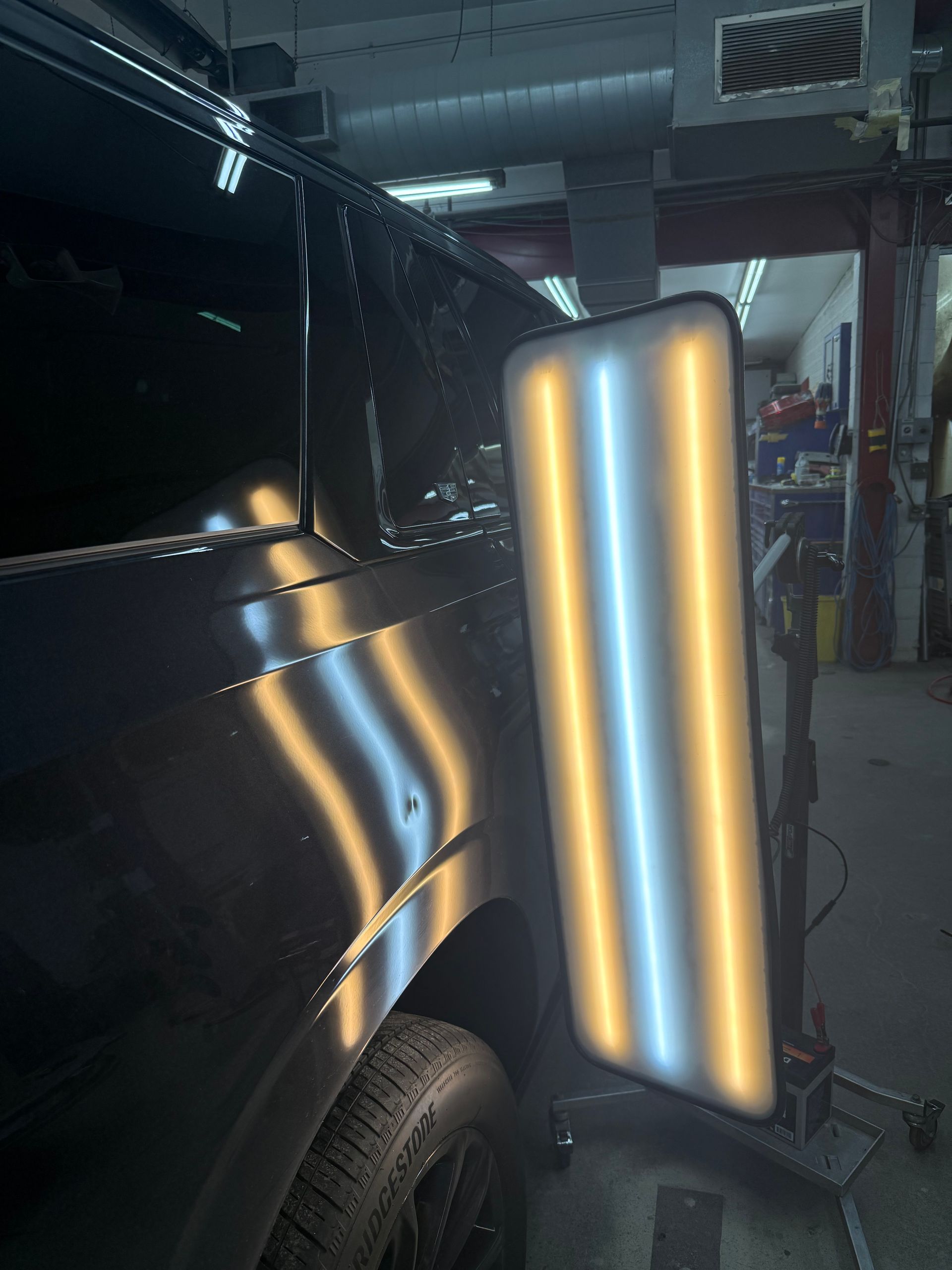 A paintless dent repair light board reflects onto a dented black vehicle panel in an auto repair shop.