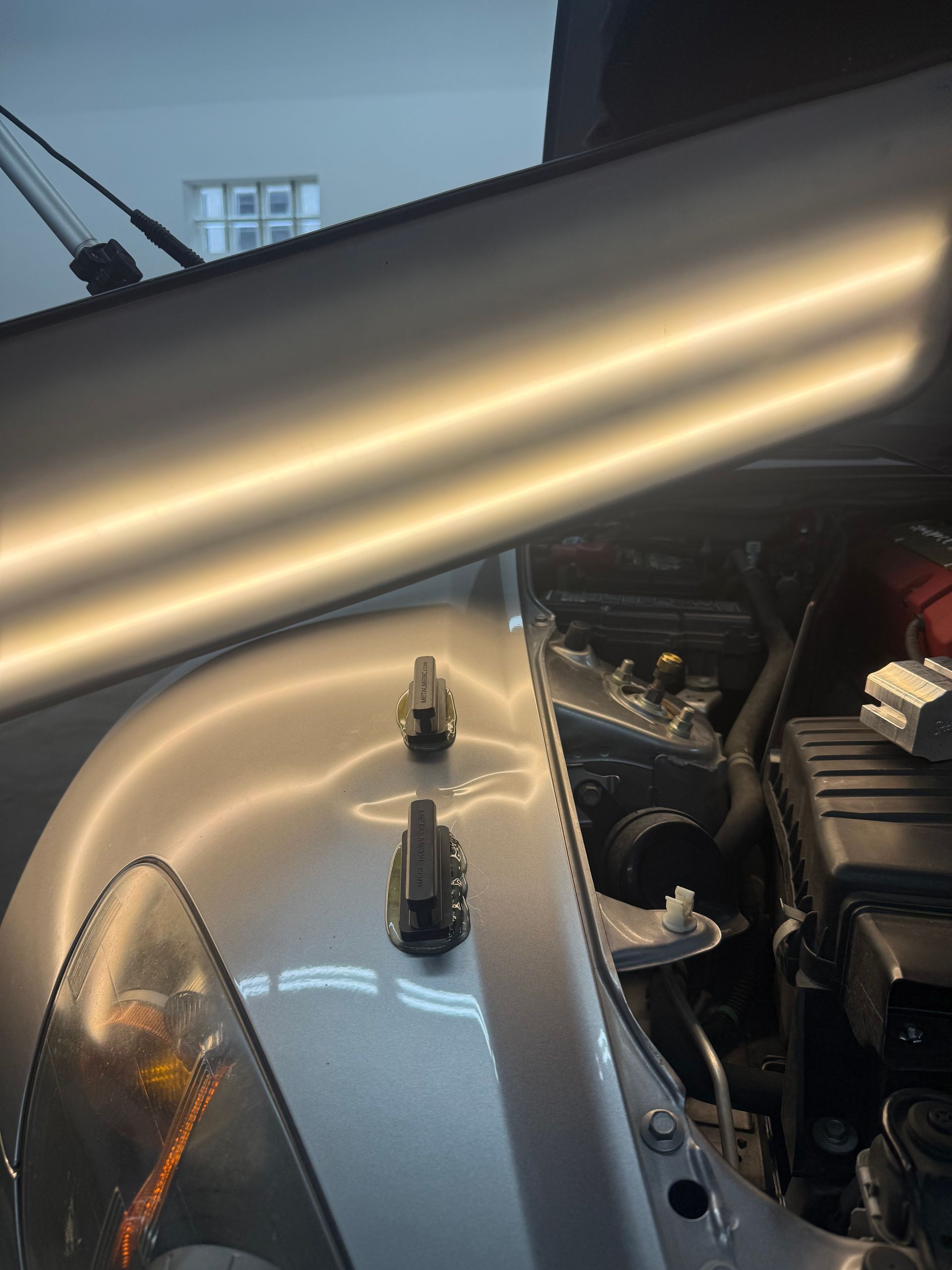 Two glue-pulling tabs are attached to a silver car fender under a shop light to repair a dent.