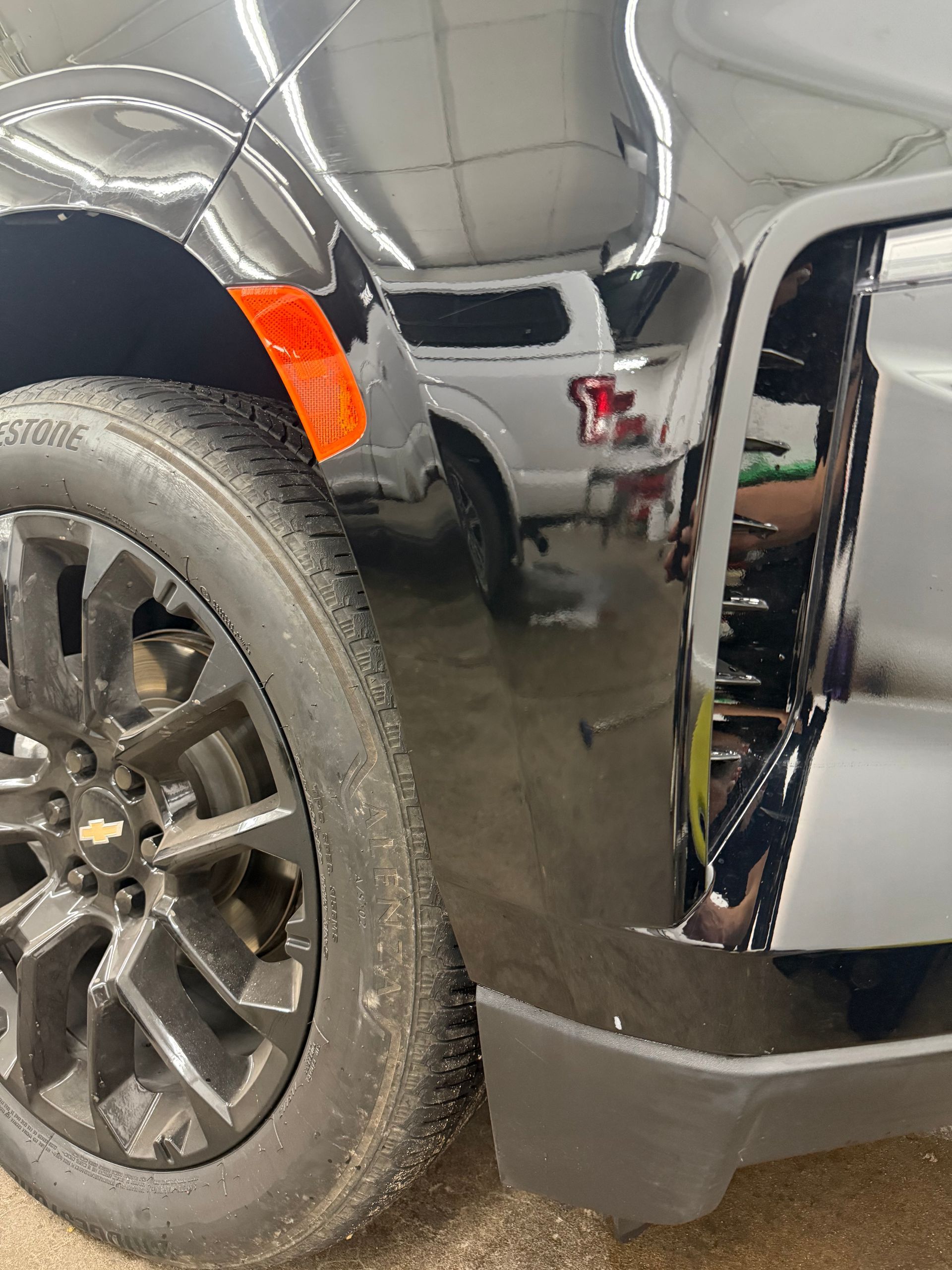 Close-up of a black truck's front wheel, orange side marker, and glossy fender reflecting a white pickup truck nearby.
