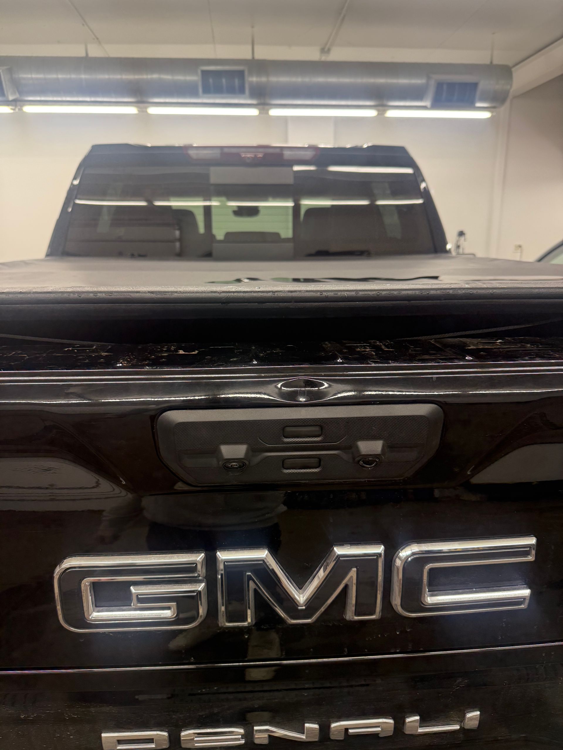 Rear view of a black GMC Denali pickup truck tailgate with a closed tonneau cover inside a garage.