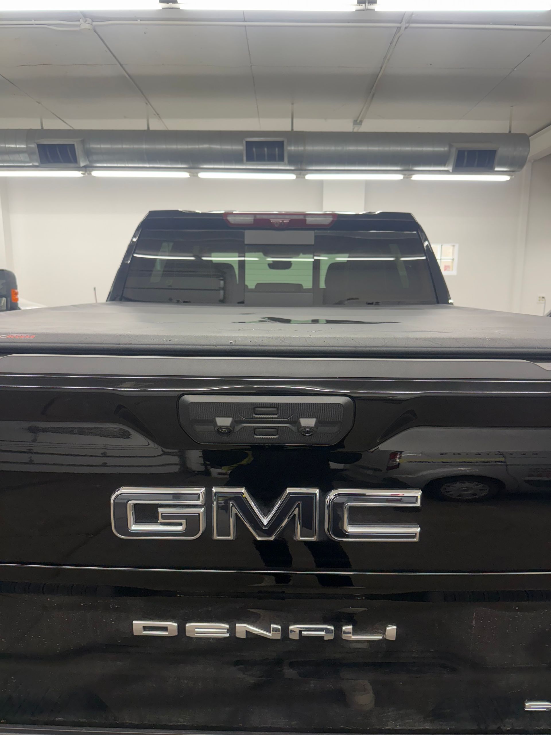 A close-up of a black GMC Denali pickup truck tailgate with a tonneau cover, centered in a brightly lit indoor garage.