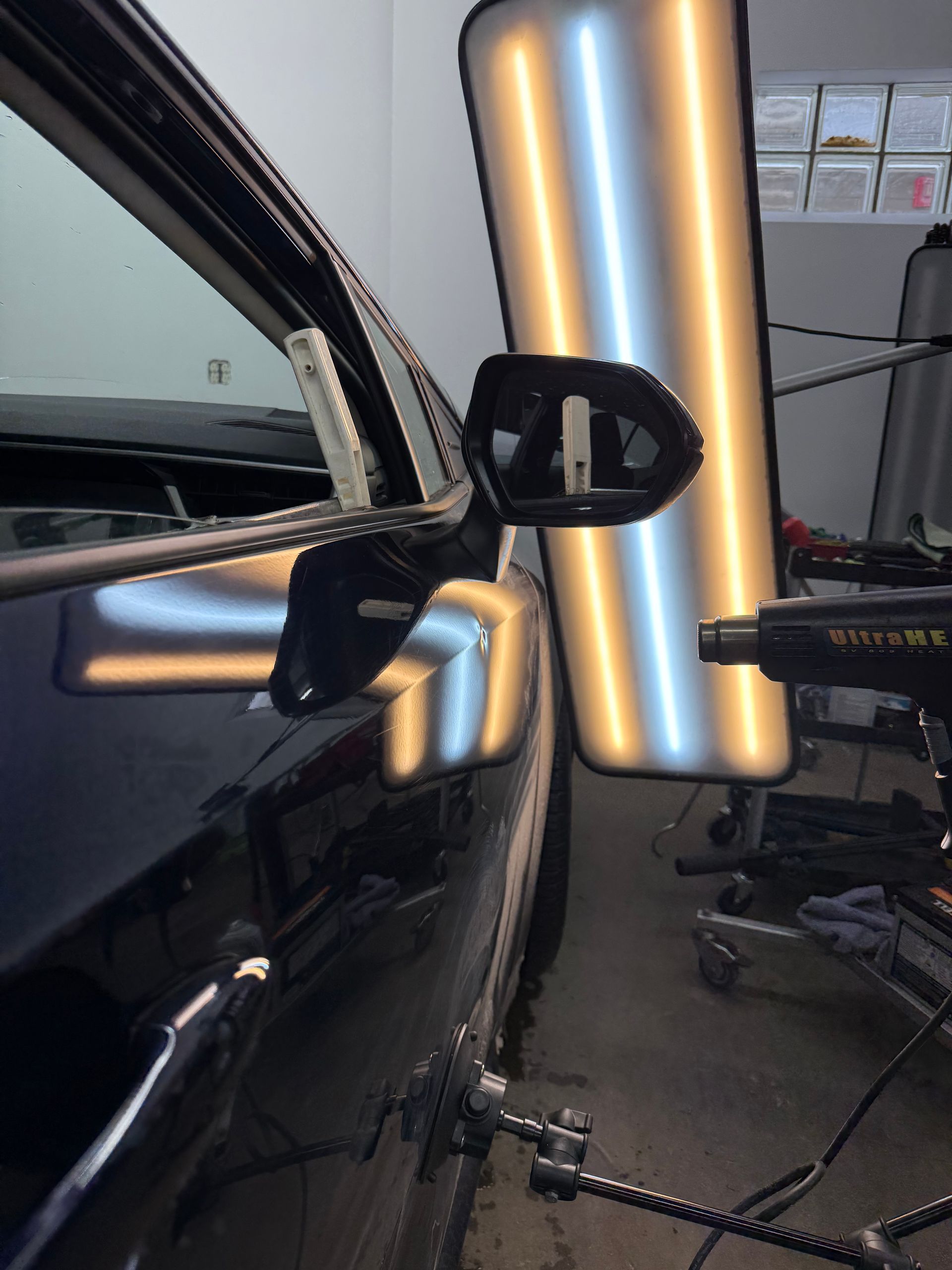 A PDR light board reflects on a black car door, assisting a technician in repairing a dent near the side mirror.