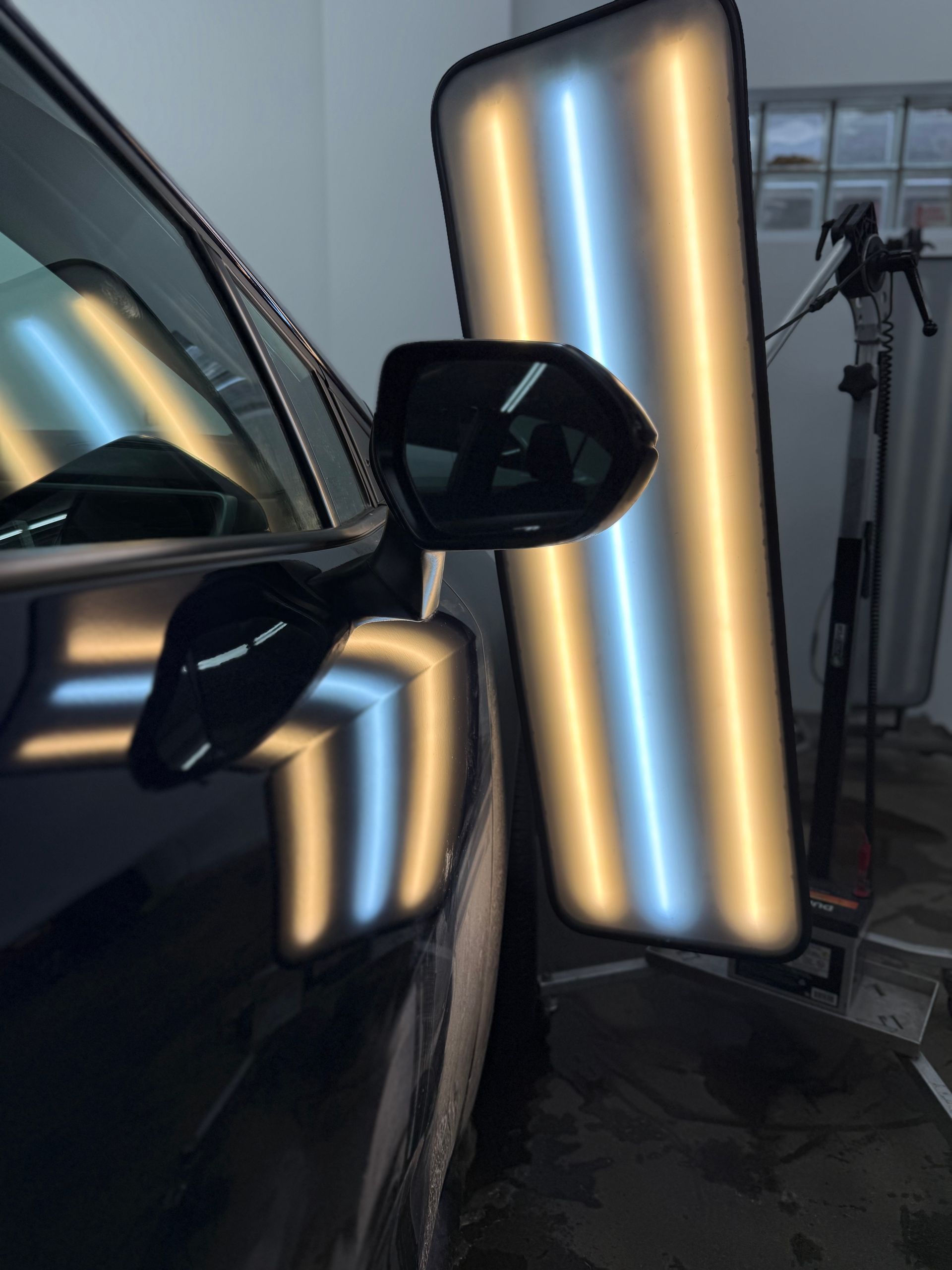 A PDR light board reflects on a car door near the side mirror, showing the panel's contour during paintless dent repair.