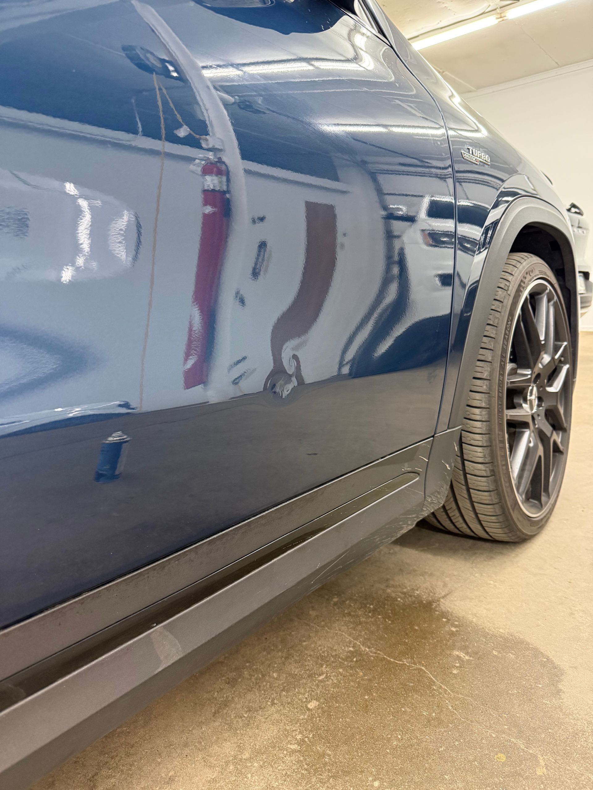 A close-up, low-angle view of a dark blue car, focusing on the passenger side door, rocker panel, and black alloy wheel.