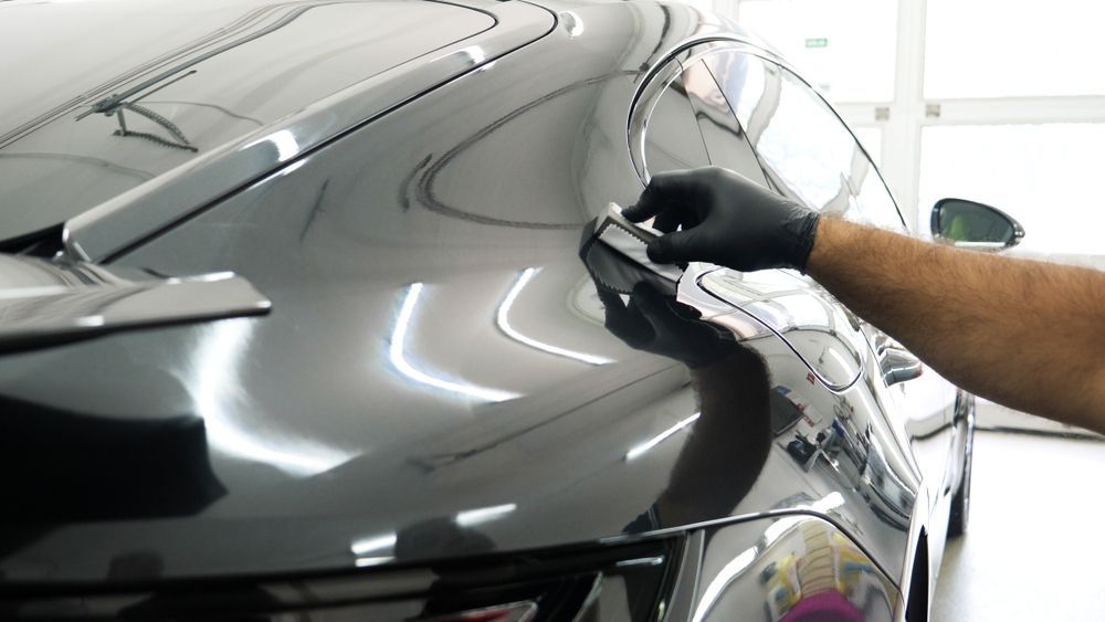 A hand in a black glove applies a ceramic coating to the glossy dark paint of a car.