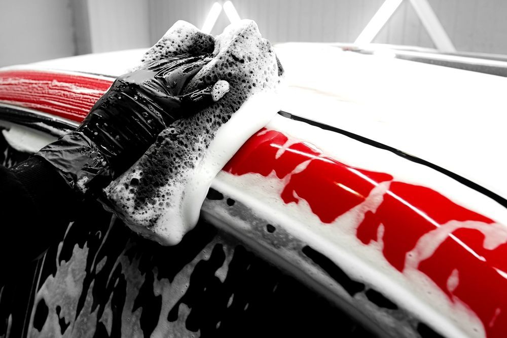 A gloved hand uses a sponge to wash a red car frame covered in white soap suds.