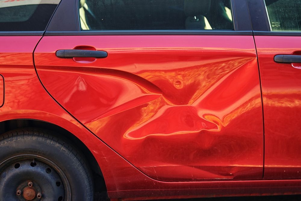 A side view of a red car with a large, concave dent across the rear passenger door.