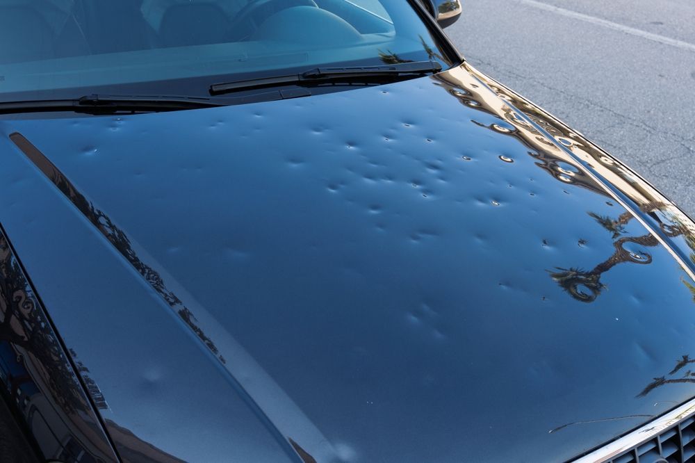 A dark-colored car hood covered in numerous circular dents caused by hail damage.