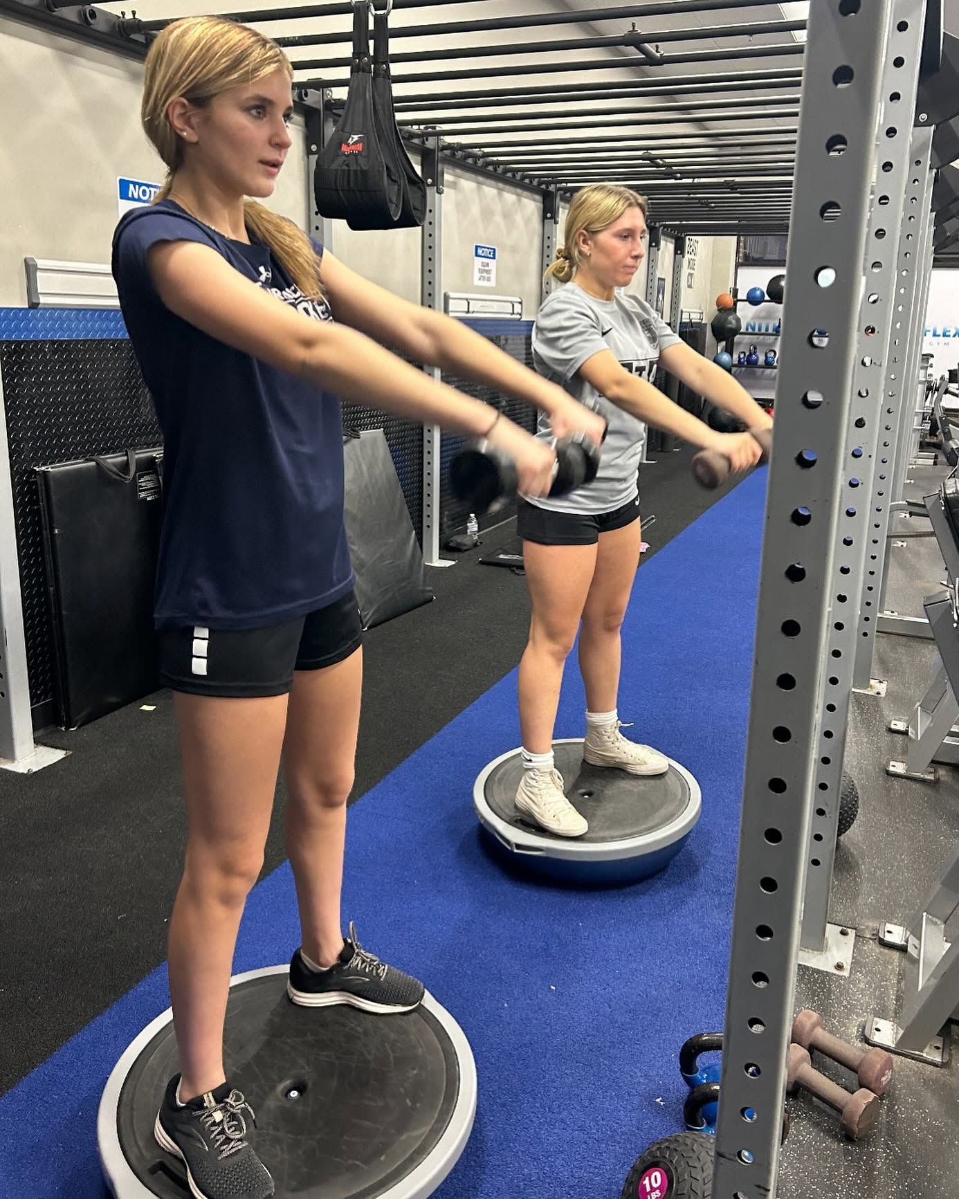 Two people balancing on wobble boards in a gym, holding arms forward for exercise.