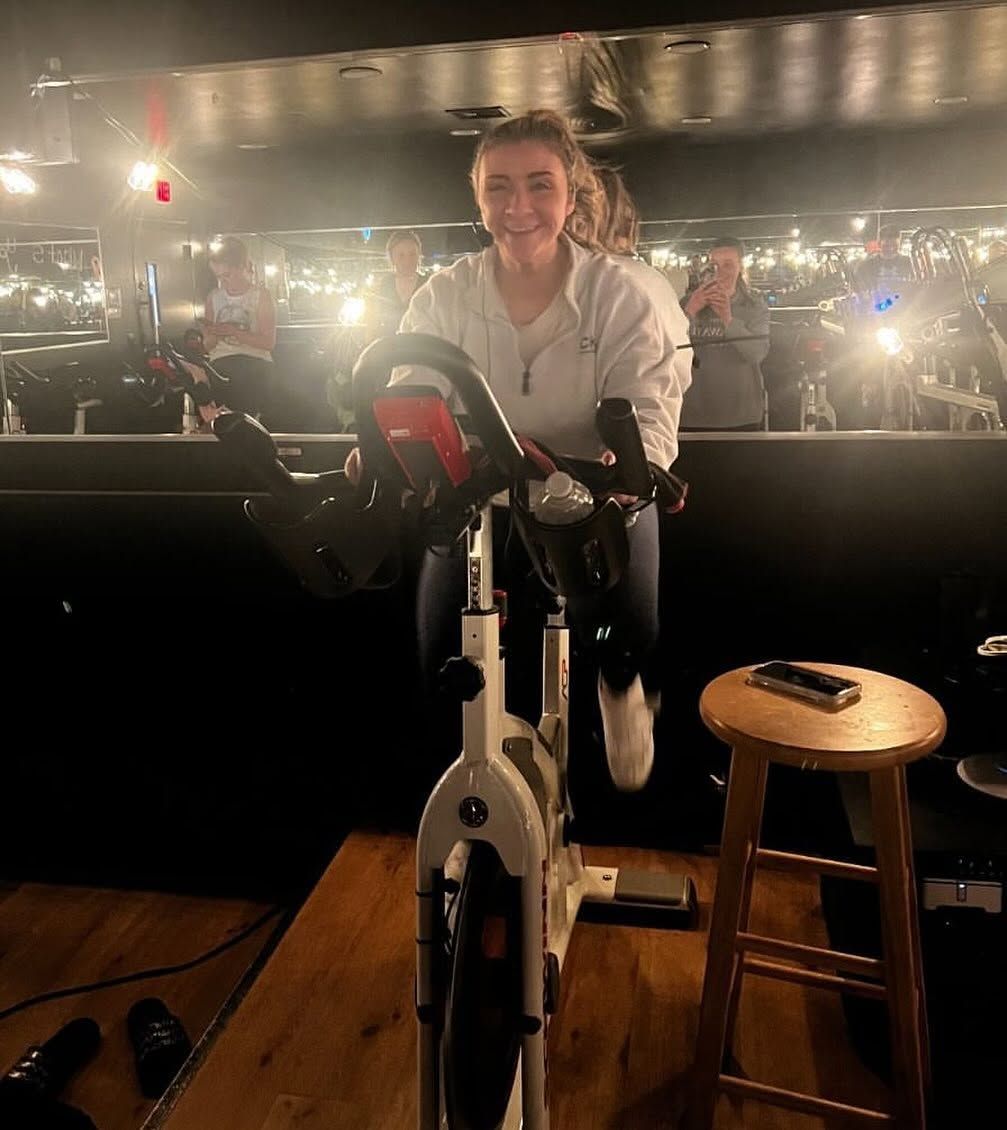 Woman smiling on a stationary bike in a dimly lit bar, with a stool beside her.