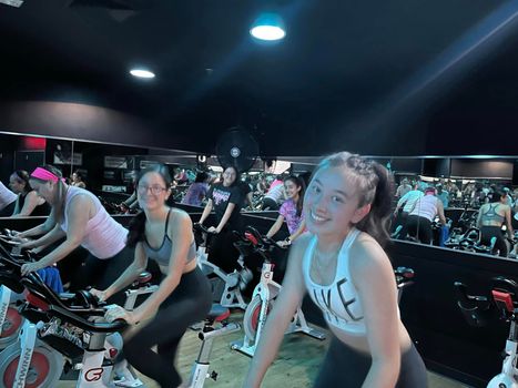People riding stationary bikes in a dimly lit indoor spin class, smiling and exercising.