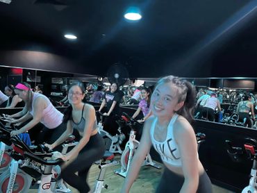 People riding stationary bikes in a dimly lit indoor spin class, smiling and exercising.