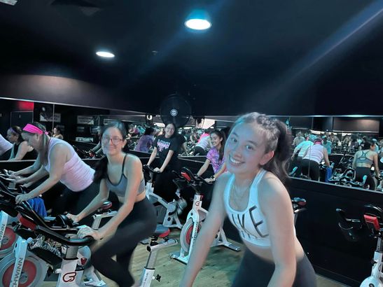 People riding stationary bikes in a dimly lit indoor spin class, smiling and exercising.