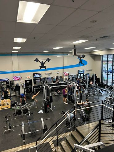 Busy gym interior with weight machines, treadmills, mirrors, and overhead fluorescent lights.