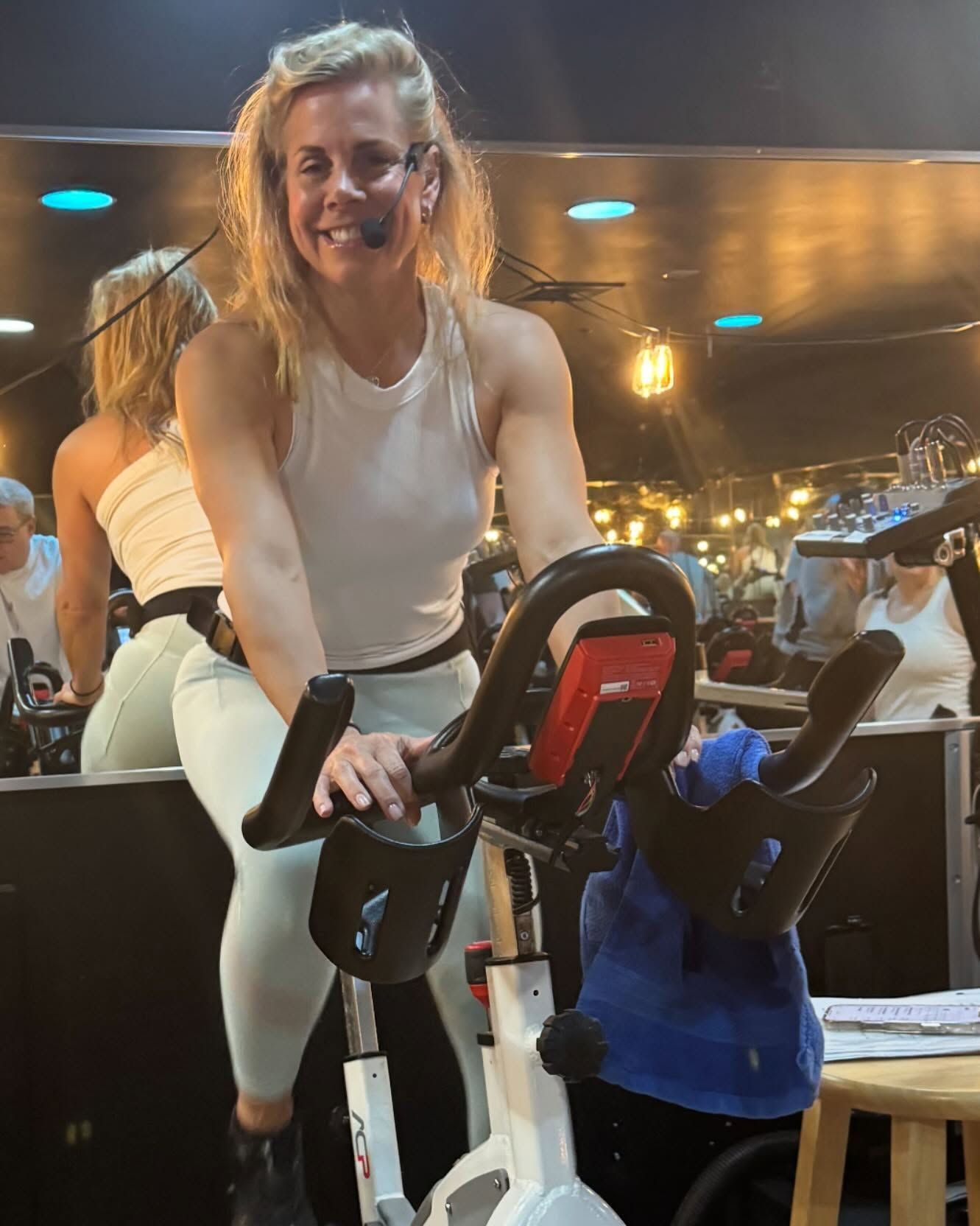 Woman smiling on a stationary bike in a gym, with reflections and warm lights in the background