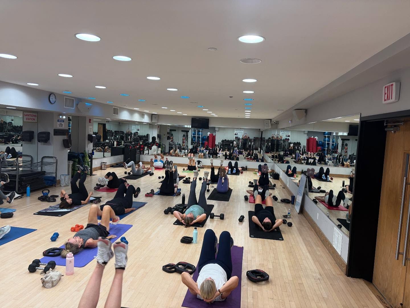 People exercising on mats in a bright mirrored fitness studio with wooden floors